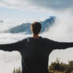 Mountain Trekking - Back view of anonymous male traveler in casual clothes standing on edge of cliff and admiring breathtaking scenery of clouds and mountain top