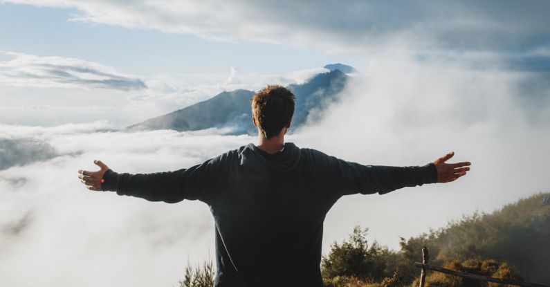 Mountain Trekking - Back view of anonymous male traveler in casual clothes standing on edge of cliff and admiring breathtaking scenery of clouds and mountain top