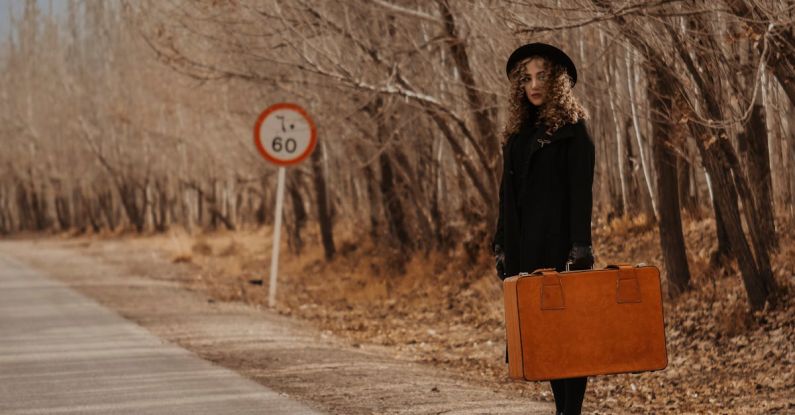 Hitchhike - Full body of female in elegant outfit and hat with old suitcase near leafless trees in autumn